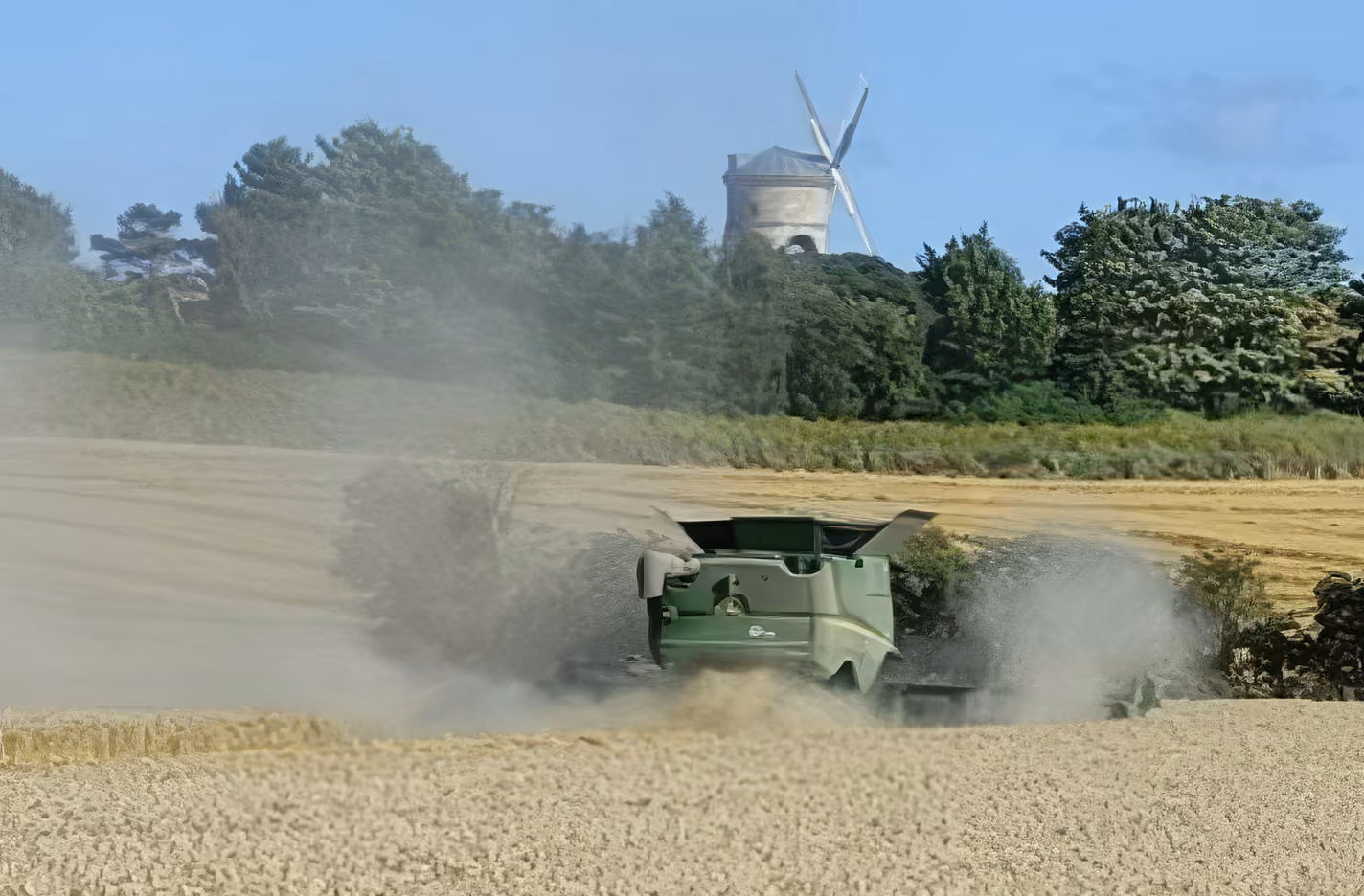 Green combine harvester working in a dusty field with trees and a windmill in the background.